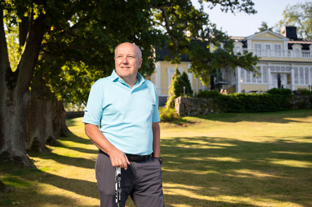 Antti Tiitola seisoo aurinkoisella nurmikolla suuren puun varjossa putteriin nojaten. Taustalla on Master Golf Clubin klubitalo, joka on keltainen kaksikerroksinen puurakennus.